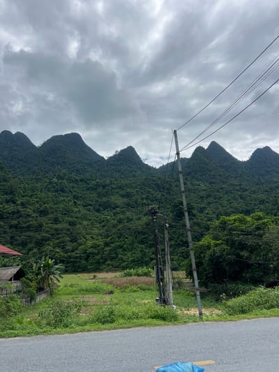 The image depicts a rural landscape in Thuận Hòa Commune, Vietnam, characterized by lush green mountains under a cloudy sky. In the foreground, a paved road runs horizontally across the bottom of the frame. To the left of the road, a small, rustic dwelling with a red-tiled roof and walls made of wood and bamboo is visible. Adjacent to the house, banana trees and other vegetation grow. A field with cultivated crops and patches of dry earth lies behind the house. In the mid-ground, two utility poles with wires traverse the scene, one more prominently to the right. The background is dominated by a range of densely forested mountains with distinct peaks. The overall atmosphere is one of tranquility and natural beauty, with a touch of human settlement. There are no people visible in the image.