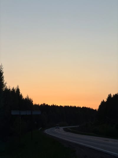 A landscape oriented photo depicts a rural setting near Oreshniki, Russia. The sky transitions from light blue to orange near the horizon. A dense forest line divides the sky from a roadway that curves to the right. A sign structure is present at the bottom left of the forest area. On the road, a single vehicle with illuminated tail lights is driving along the road.