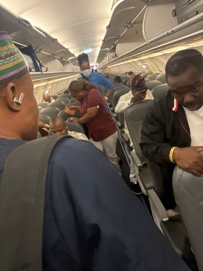 The image shows the interior of an airplane cabin, presumably in flight. Passengers are seated in rows along the aisle. Individuals are visible in varying degrees of focus and orientation, suggesting a candid, perhaps spontaneous, capture of the scene. One person in the foreground is wearing a patterned hat and has a visible earring. Further down the aisle, other passengers are engaged in various activities. The location in Omoecheigbo, Nigeria is not readily identifiable from the visual content of the picture; the setting is identifiable as an airplane interior based on the structural elements and configuration of seating.