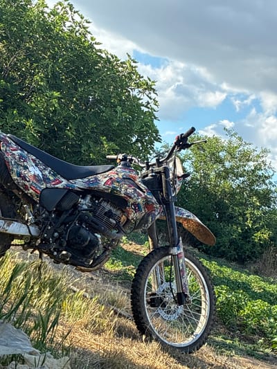 A dirt bike with a colorful, graffiti-style decal wrap is parked on a grassy, uneven terrain with dry grass and some green foliage. The bike is angled towards the viewer's left, with its front wheel prominently displayed. The background consists of lush green trees and a cloudy blue sky. The scene appears to be outdoors, possibly in a rural area outside of Glufishevo, Bulgaria, given the natural vegetation and the absence of urban structures. There are no people or any actions depicted in the image.