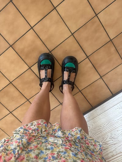 The image shows a person's legs and feet, viewed from above. The person is wearing black platform Mary Jane shoes with multiple straps and silver buckles. A green insert is visible inside each shoe. The person is also wearing a short, floral-patterned dress. The floor is tiled with large, square, light brown tiles, and there is a section of light-colored wooden planks to the right of the tiles. The image does not contain any information that would allow for the determination of a specific location in the city of Nivigne et Suran, France.