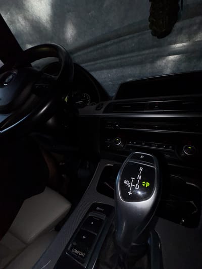 The image depicts the interior of a car, specifically the dashboard area. The focal point is the gear shifter, which is illuminated and displays various driving modes including Park (P), Reverse (R), Neutral (N), Drive (D), and Manual/Sport (M/S). The steering wheel is visible on the left side of the image. Buttons and controls for the car's climate system and infotainment are also present on the dashboard. The overall lighting suggests it is either nighttime or the car is in a dimly lit environment. The car appears to be parked, as indicated by the 'P' on the gear shifter. There is no discernible indication of the car's location in Novy, Russia.
