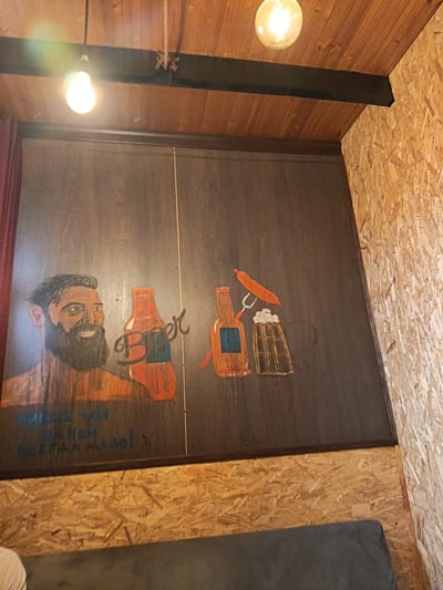 This media file captures a portion of an interior space, likely a bar or cafe, with a distinct rustic and unfinished aesthetic. The main subject is a dark, wooden panel on which a mural-like drawing is displayed. On the left side of the panel, a man with a prominent beard and dark hair is depicted, looking towards the viewer with a slight smile. To his right are two orange-tinted bottles, one of which has the word "Beer" scrawled in a stylized font across its front. Further to the right, another bottle, a sausage impaled on a fork, and a frothy mug of beer are illustrated. Below the drawings, in blue lettering, are the Cyrillic words "Пиво и Чай" (Beer and Tea) and "Всегда мало!" (Always not enough!). The background of the room features exposed wooden beams on the ceiling and walls constructed from oriented strand board (OSB), giving it a raw and perhaps unfinished feel. Two light bulbs hang from the ceiling, one a bare incandescent bulb and the other a warmer, Edison-style bulb, suggesting it is likely evening or interior lighting is the primary illumination. The overall impression is of a casual, perhaps slightly quirky, establishment in Kasumkent, Russia, decorated with playful artwork related to beverages and snacks.