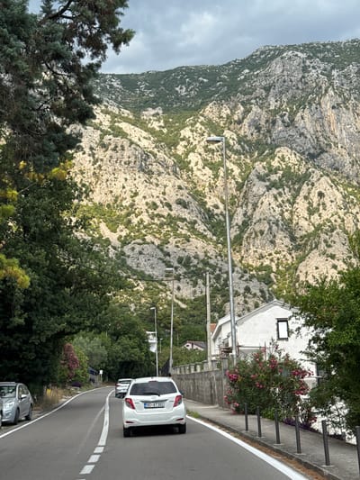 The image shows a road winding through a mountainous area in Donji Orahovac, Montenegro. In the foreground, a white Toyota Yaris is driving on the road, followed by another car. The road is lined with trees and bushes, with a white building on the right side of the road. In the background, a steep, rocky mountain rises, covered with sparse vegetation. The sky is overcast, and the lighting suggests it is daytime. The scene appears to be a scenic route, possibly on the coast of the Bay of Kotor.