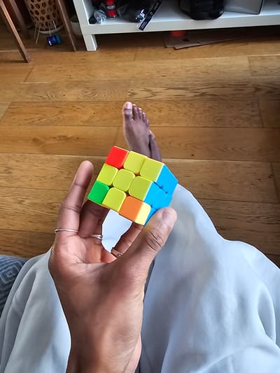 A person is holding a Rubik's Cube. The cube has colors blue, yellow, orange, red, and green. The person's hand is holding the cube in front of a light blue or white garment. The background is a wooden floor. Part of a white shelving unit is visible on the left. The location in Pantin, France is not determinable from the image.