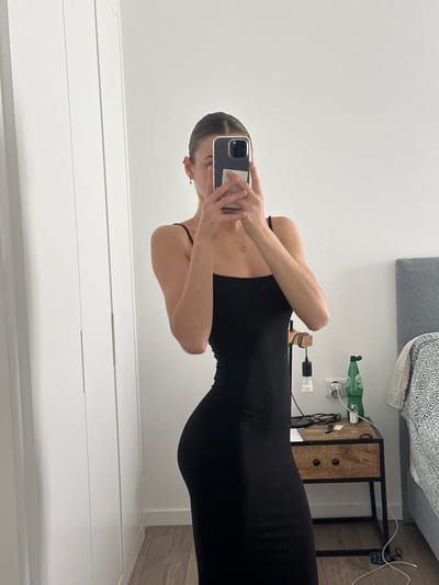 The image depicts an indoor mirror selfie, presumably taken in a bedroom located in Ericeira, Portugal. A female subject is captured from the mid-chest down, wearing a form-fitting black spaghetti-strap maxi dress. Her face is obscured by the dark-colored smartphone she holds directly in front of her to capture the reflection. She wears small earrings and a delicate necklace with a circular pendant. To the left, a white wardrobe or cabinet with vertical paneling is visible. To the right, a wooden nightstand with two dark drawer pulls and a dark metal frame stands next to a bed with a grey headboard and a patterned blanket. On the nightstand, a small wooden lamp with an exposed bulb hangs, alongside a green glass bottle, a white power outlet with a visible plug, and several small dark objects. The room features white walls and light-colored wood laminate flooring.