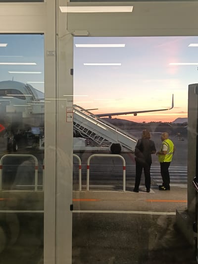This image depicts a scene at the Alghero Airport in Alghero, Italy, at dusk. Through large glass doors, an airplane is visible on the tarmac, partially obscured by a jet bridge. Two individuals are standing outside the doors, facing the aircraft. One, a person with long brown hair, wears dark clothing. The other, a man, is dressed in a high-visibility yellow vest over dark clothing. A set of mobile stairs is positioned next to the airplane. The sky in the background displays the soft colors of sunset. The airport environment is suggested by the tarmac, markings, and equipment.