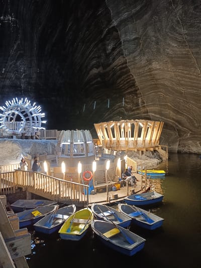 The image shows the interior of the Turda Salt Mine in Turda, Romania. The scene depicts an underground lake with several rowboats floating on its surface. On the right side of the lake, there is a wooden structure built on stilts with a roof made of angled wooden beams. On the left side, a large, circular, illuminated structure resembling a wheel with multiple light bulbs is visible. A wooden walkway with railings surrounds part of the lake, connecting different structures and offering access to the boats. Several people are seen on the walkway and near the wooden structure, some appearing to be tourists. The walls of the mine are rough and textured, with visible striations characteristic of salt formations, illuminated by artificial lights, creating a dramatic, cavernous atmosphere. The water is dark and reflects the surrounding lights.
