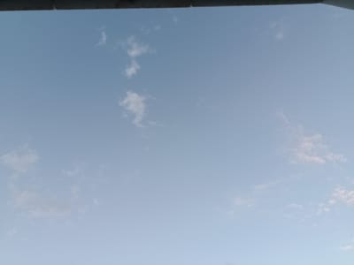 The image shows a light blue sky with sparse, wispy white clouds. A dark, horizontal bar runs across the top of the frame, obscuring the very top of the sky. The sky appears to be clear and vast. The location in the city of Arkadag, Turkmenistan, is a residential area or a public space with a clear view of the sky. There are no people or specific objects clearly visible in the image besides the sky and clouds, and the dark horizontal bar at the top which could be part of a building or structure. The scene is calm and uneventful.