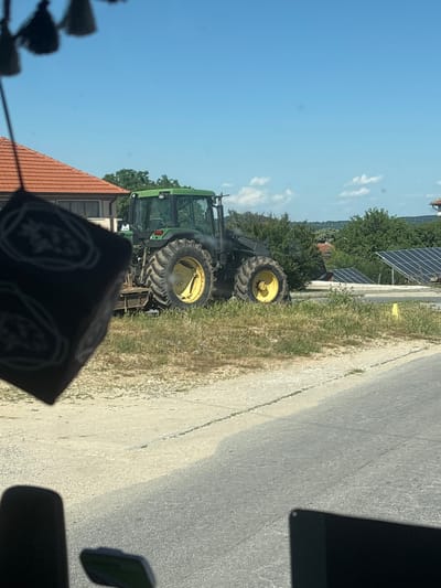 An outdoor daytime shot shows a green tractor with large yellow wheels parked on a gravel and grass area. The tractor is facing to the right. To the left of the tractor is a building with a red-tiled roof. In the background, there are trees and a field of solar panels. The foreground shows a section of a road. The image is partially obscured by a dark object hanging from above on the left side, and by parts of a vehicle interior at the bottom. The location is specified as Hliabovo, Bulgaria.