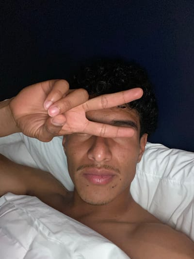 A person lies in bed with a white pillow and comforter. They are making a peace sign with their right hand, which is held up close to their face. The person has dark curly hair and a slight mustache. They are shirtless and appear to be lying on their back. A dark blue fabric or surface is visible behind their head.