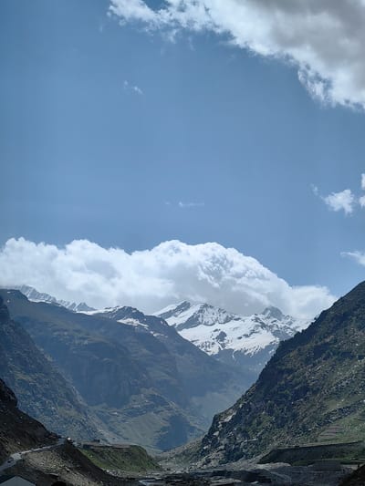 The image depicts a mountain landscape in the vicinity of Burwa, India. The scene is dominated by towering, rugged mountains, some of which are capped with snow. In the foreground, a winding road snakes through the valley, with several vehicles visible, suggesting it is a route for transportation. The slopes of the mountains are covered in sparse vegetation and rocky terrain. A river or stream appears to flow through the valley floor. The sky above is a clear blue with scattered white clouds, some of which are large and billowy, obscuring parts of the mountain peaks. The overall impression is one of a remote, mountainous region with signs of human activity in the form of a road and vehicles.