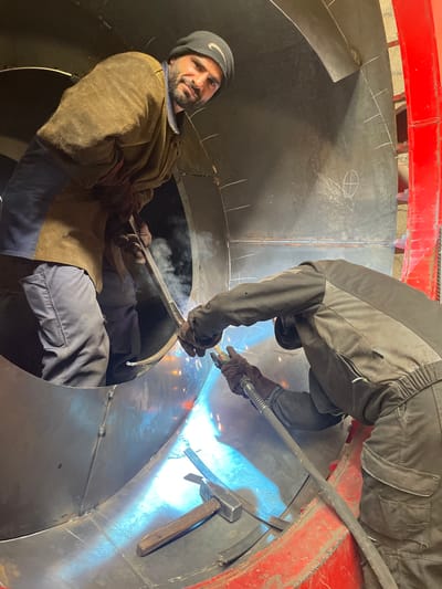 Two welders are working inside a large, curved metal structure, likely a section of a pipe or vessel. One welder is kneeling and welding a seam, while the other is positioned above, also welding. Sparks and smoke are visible from the welding process. A hammer rests near the welding area. The location, Tomba, Montenegro, is not discernible from the visual information provided.