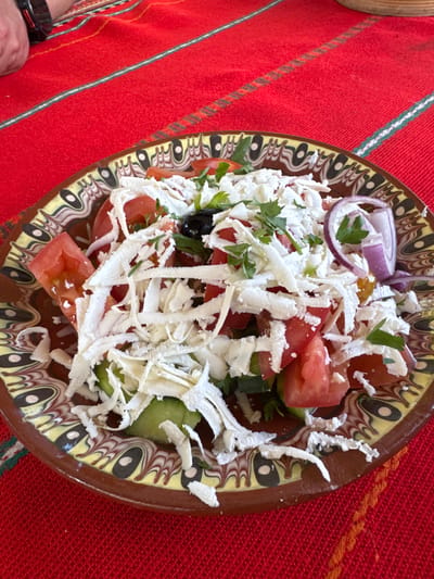A colorful ceramic plate containing a Bulgarian Shopska salad rests on a red and white patterned tablecloth. The salad consists of diced tomatoes, cucumbers, red onion, parsley, black olives, and shredded white cheese (likely sirene). The scene is located in Kalimantsi, Bulgaria. A portion of a wrist with a watch is visible at the bottom left.