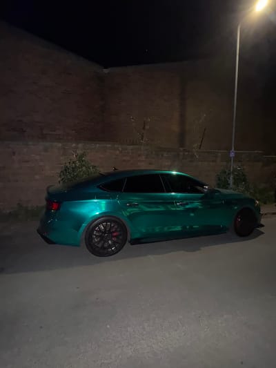 A teal or metallic emerald green car, likely a sporty sedan or hatchback, is parked outdoors at night in South Kesteven, United Kingdom. The image is rotated 90 degrees to the left. The car features black multi-spoke alloy wheels with red brake calipers visible through the spokes. A bright street lamp, serving as the main light source, is positioned in the upper left of the frame, casting illumination on the car and its immediate surroundings.
To the left of the car, a dark brick wall with visible texture can be seen. Behind the car, sparse, dark green foliage or bushes are present. The ground beneath the car appears to be a dark, paved surface like asphalt or concrete. On the right side of the car, a lighter-colored wall or surface, possibly concrete, is visible. No people are present, and no specific activity is taking place other than the car being parked. The scene is characterized by its nighttime setting and the artificial light from the street lamp. No discernible text is visible on the car or in the environment.