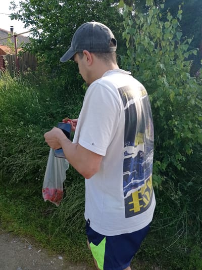 A young man wearing a gray baseball cap, a white t-shirt with a graphic print, and navy blue shorts with lime green accents is standing outdoors near a dirt path. He is holding a clear plastic bag filled with red strawberries in his left hand, and a smartphone in his right hand, appearing to be taking a picture of the berries. He is looking down at the strawberries and phone. The background consists of lush green foliage and trees, with a portion of a wooden fence and a building visible to the left. The scene suggests a rural or suburban environment, potentially within the city of Degtyarsk, Russia, given the context provided.