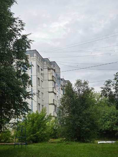 The image depicts an overcast day in Mishutino, Russia, with a residential apartment building as the central focus. The building, constructed with white and light blue tiled panels, features multiple stories with visible windows and balconies. Several satellite dishes are affixed to the exterior. Surrounding the building is a grassy area with various trees and bushes in different shades of green. A blue metal playground structure is visible in the foreground on the left side of the image. Overhead, power lines crisscross the sky, and several birds are in flight. In the lower right corner of the frame, a white object, possibly a piece of discarded material, lies partially hidden in the grass.