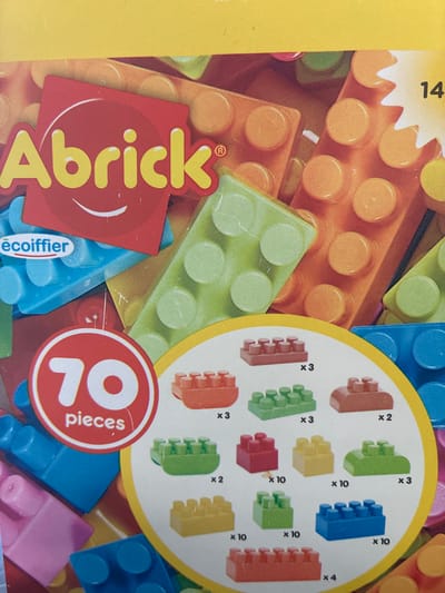 This media file is a close-up, top-down photograph of a toy box. The box is filled with colorful building blocks, primarily in shades of orange, green, blue, and red, with some yellow and pink pieces visible. The blocks appear to be plastic and are of various shapes and sizes, including rectangular bricks with raised studs and smaller, rounded pieces.
Dominating the foreground is a red sticker or label with the word "Abrick®" in large yellow, stylized letters. To the right of this, a circular red emblem features the number "70" in white, with the word "pieces" also in white written vertically beside it. Another smaller oval label with the text "écoiffier" in white is partially visible on a blue block.
The background is a blurry assortment of more building blocks, suggesting a large quantity of toys. Some text is visible on the box itself, including the number "14" in black on a yellow background at the top left, and smaller text labels with "x3", "x10", "x2", and "x4" next to images of different block configurations, indicating the quantity of each type of block. The lighting is bright, and the overall impression is of a vibrant, child-oriented product. There are no people or animals visible, and the setting is contained within the packaging of the toy. The scene is likely indoors, given the artificial lighting and the nature of the product. The image does not provide any clues about the time of day, weather, or specific emotions, beyond the playful and exciting nature of toys. The location context is Toruń, Poland, but this cannot be discerned from the image itself.