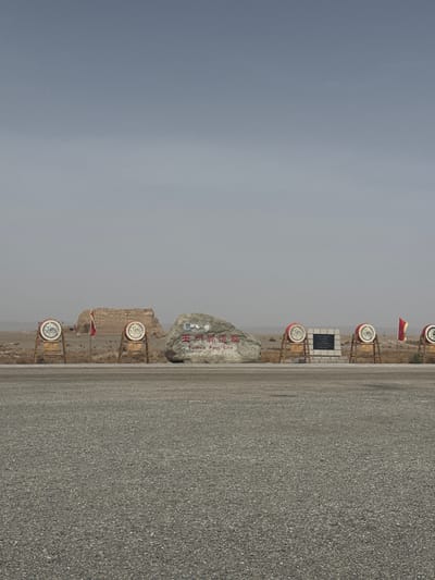 The image depicts a stark, open-air scene, likely an entrance or landmark at a historical site. The perspective appears to be slightly rotated, but if oriented correctly, a wide, granular road or pathway would occupy the lower right portion of the frame, stretching into the background.

To the left of this road, a prominent, light-colored natural stone boulder stands, bearing the site's name in both Chinese and English. Clearly visible on the stone are the Chinese characters "玉門關" (Yumen Pass) and the English text "Yumen Pass." Near the top left of the stone, some small, unclear symbols or characters are also present.

Extending along what appears to be a parallel path or demarcation, several distinct circular signs are mounted on wooden posts. These signs feature red tops and white centers, which may contain decorative symbols or motifs, though they are not clearly discernible. A small red flag on a pole is visible among these signs, adding a touch of color to the muted palette. Further back, a dark, rectangular plaque is mounted on a stone base, but any text on it is too small to read.

The background reveals an arid, desolate landscape characterized by low, rocky formations or hills, typical of a desert environment. The sky is a uniform, hazy light grey, suggesting an overcast day or significant atmospheric dust, obscuring direct sunlight. The overall color palette is dominated by muted greys, browns, and beiges, with the red and white elements of the signs and flag providing minor contrasts.

The scene is static, devoid of any human activity. The presence of "Yumen Pass" text directly indicates the location, a historically significant pass associated with Dunhuang City, China.