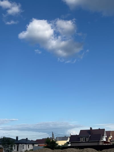 A low-angle, vertical photograph captures a bright blue sky with scattered white and gray clouds. In the upper portion of the frame, a large, fluffy white cloud dominates the center. The lower third of the image features the rooftops of several houses, suggesting a residential area. The architecture appears to be modern, with dark-colored roofs and light-colored walls. Some houses have visible chimneys and dormer windows. Overhead power lines are also present, stretching horizontally across the frame. In the foreground, mounds of dirt or sand are visible, indicating ongoing construction or landscaping. The scene is consistent with a suburban or rural environment, possibly in Novaya Usman, Russia, given the architectural style and the presence of power lines. No people are visible in the image.