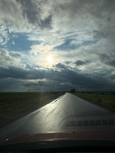 A road stretches straight ahead under a sky filled with dynamic clouds. The sun shines brightly through a break in the cloud cover. To the right of the road, part of a vehicle interior is visible, suggesting the photo was taken from inside a car. Green fields line the road. The image provides no specific clues regarding its location in Verkhnie Iziverki, Russia.