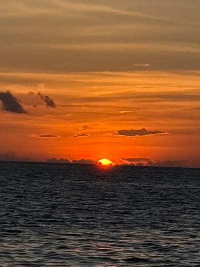 The image depicts a sunset over the ocean in Baan Taling Ngam, Thailand. The sky is filled with vibrant orange and yellow hues, with streaks of darker orange clouds scattered across. The sun is low on the horizon, partially obscured by a thin layer of clouds, casting a warm glow over the scene. The ocean is dark and choppy, reflecting the colors of the sky. The horizon line is clearly visible, separating the sky from the sea. There are no people or man-made objects visible in the image.