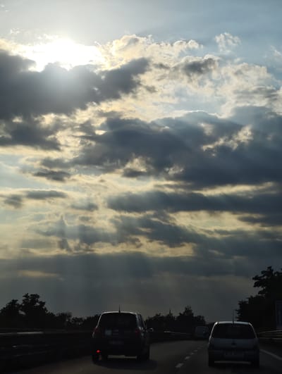 The image shows a view from a vehicle traveling on a highway in Acerra, Italy. The sky is filled with dramatic clouds, with the sun breaking through in the upper left, casting beams of light downward. Several vehicles are visible on the road, including the back of a dark-colored car in the foreground, a light-colored car in the right lane, and another vehicle further ahead. Trees line the sides of the highway, silhouetted against the brightly lit sky. The scene suggests a late afternoon or early evening with an approaching storm or the aftermath of one.