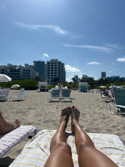 The image shows a person lying on a beach lounger on South Beach in Miami Beach, Florida, at the coordinates 25.769, -80.132. The person's legs are visible in the foreground, with their feet resting on a striped towel. The beach is sandy and there are several other loungers visible in the background. There are also several buildings in the background, as well as people walking on the beach. The sky is clear and blue. The scene is a typical sunny day on South Beach, Miami Beach.