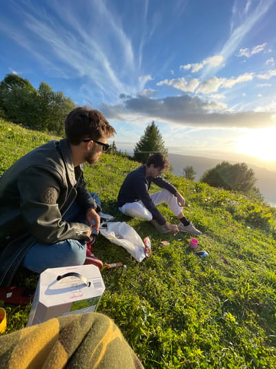The image depicts two males seated on a grassy hillside in Les Déserts, France, under a late afternoon sky. The male on the left wears a dark leather jacket, blue jeans, and dark sunglasses. He has dark hair and a beard, and is seated with legs crossed, looking towards the right side of the frame. The male on the right wears a dark long-sleeved shirt, light-colored trousers, and grey sneakers. He has dark, curly hair, is leaning forward, and is in the process of lighting a cigarette with a silver lighter. A white cardboard box, labeled "Les Ormes Cambras Grenache", is positioned in the immediate foreground, alongside a red-cased mobile phone. A crumpled white plastic bag, an orange packet marked "OCB", and a pink plastic cup are scattered on the grass near the individuals. A yellow and dark-patterned blanket is visible in the bottom-center foreground. The setting features a vibrant green, gently sloping grassy terrain dotted with small wildflowers and scattered deciduous and coniferous trees. The sky is bright blue with elongated cirrus clouds and lower, darker cumulus clouds near the horizon. The sun, low on the right, casts warm, golden light across the distant mountains.