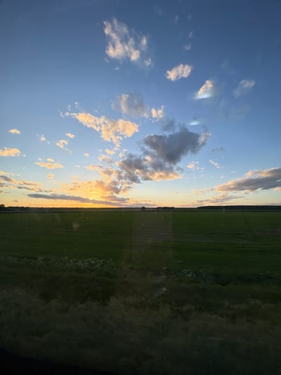 The image depicts a landscape in Мажэйкаўскі сельскі Савет, Belarus, split between a sky and a field. The sky, primarily blue, is populated with scattered, sunlit clouds, particularly dense towards the horizon. The field appears green and stretches towards the horizon, bordered by a line of trees. The sunset casts a golden hue on the clouds and the horizon.