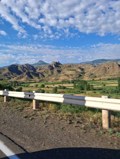 The image captures a scenic view of rolling hills and mountains under a partly cloudy blue sky, likely taken from a moving vehicle on a road. In the foreground, a white guardrail with concrete posts separates the road from the landscape. The road surface appears to be asphalt with a white lane marker visible on the left. The landscape beyond the guardrail features cultivated fields of varying shades of green and yellow, interspersed with trees and shrubs. In the background, rugged, arid hills rise up, with a distinctive conical mountain peak visible further in the distance. The overall scene suggests a rural or semi-rural setting, possibly in the Vayots Dzor Province of Armenia, given the mention of Yeghegnadzor. There are no people or specific actions depicted in the image. The lighting indicates it is daytime, with the sun casting shadows.