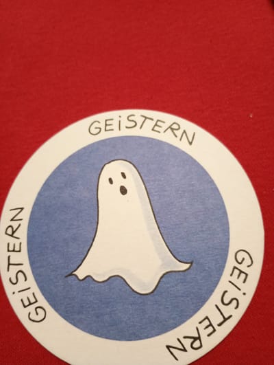 A circular sticker with a white border and a blue background is placed on a red fabric surface. The sticker depicts a cartoon ghost in the center. The ghost is white with two black dots for eyes and a black oval for a mouth. The word "GEISTERN" is written in black text around the outer edge of the sticker, split between the top and the bottom arcs. The sticker appears to be a novelty item or a decorative element. The red fabric is textured and appears to be a close-up shot, suggesting the sticker is a part of a larger item or surface. No specific location in Rosenheim, Germany, can be identified from the image.