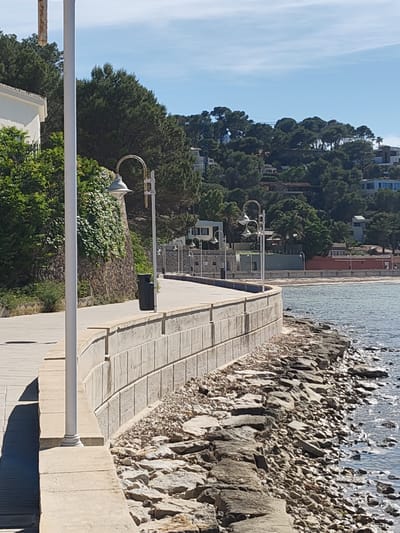 The image shows a coastal scene, likely a promenade in Rotes, Spain. A curved, low concrete wall separates a paved walkway from a rocky shoreline and the sea. Several evenly spaced, vintage-style lampposts line the walkway. The area beyond the shoreline is a mix of trees and buildings, suggesting a residential or resort area on a hillside. The sky is blue with some light clouds. A tall, white post is prominently positioned on the left side of the photo, partially obstructing the view. A black trash receptacle is visible near the wall. There are no people evident in the photo.