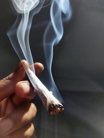 An individual's hand, displaying a darker skin tone, is captured in close-up, holding a lit hand-rolled smoking item. The item, appearing to be partially consumed, exhibits a burnt, reddish-brown tip and a white paper wrapper. Multiple plumes of white and light blue smoke emanate from the burning tip, rising upwards and to the right, forming distinct, swirling patterns against a uniformly dark gray, indistinct background. The scene is illuminated from the left, highlighting the hand, the smoking item, and the rising smoke. This event occurred in Ksar El Majaz, Morocco.