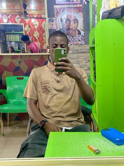 A Black male, wearing a horizontal striped brown and black polo shirt with an emblem, dark grey pants, a black digital watch, and a gold ring on his right hand, takes a mirror selfie in a barbershop or salon located in Zaki Biyem, Nigeria. He holds a dark green cased iPhone, largely obscuring his face, and wears white wireless earbuds. He is seated at a light green counter or table, on which an orange disposable lighter and a bright blue smartphone case are present. The background, visible in the mirror's reflection, includes patterned wallpaper (red and gold, and a geometric red/black/brown design), a stone-textured wall, and posters featuring Tupac Shakur and various hairstyles. In the reflection, another Black male in a red sleeveless shirt is seen attending to a customer in a barber's chair. Additional green plastic chairs, shelves stocked with white product bottles, and a ceiling fan are also visible in the reflection. A solid lime green partition or cabinet section is visible to the subject's right.