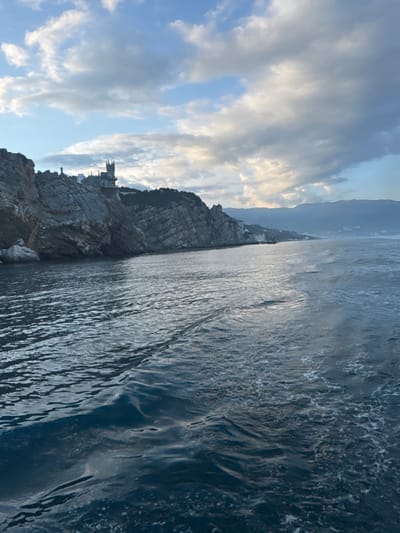 The image shows a coastline with a rocky cliff on the left and the sea on the right. At the edge of the cliff, there is a castle, presumably Swallow's Nest, a popular landmark in Gaspra, Ukraine. The sky above is filled with clouds, with patches of blue visible. The sea appears rough with reflections of the sky. In the background, there is a hazy mountainous landscape. The photograph appears to be taken from a slightly elevated position, possibly from a boat or another cliff overlooking the scene.