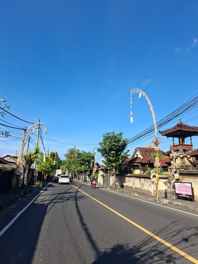 A clear blue sky is visible above a two-lane asphalt road in Banjarangkan, Indonesia. On the right side of the road, a traditional Balinese temple structure with a tiered roof is adorned with decorative poles that extend upwards and are decorated with white fabric and a yellow flag. Adjacent to the temple is a sign for "smartfren". Buildings with terracotta roofs line the left side of the road. Several trees with lush green foliage are present along the roadside. A white car is driving away from the viewer in the distance, and a red motorcycle is approaching. The road is marked with a yellow line down the center and a white line on the left edge. Shadows from overhead wires and trees are cast on the road surface.