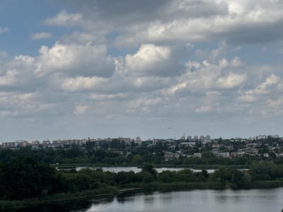 A wide shot captures a cityscape from a distance, with a body of water in the foreground. The sky is filled with a mix of blue patches and dramatic, heavy clouds.
In the middle ground, a dense line of green trees separates the water from the city. Beyond the trees, a sprawling urban area is visible, characterized by a mix of low-rise buildings and taller apartment blocks. The architecture suggests a residential and possibly industrial or commercial district. A faint airplane is visible in the sky above the city.
The foreground shows a calm, dark body of water, possibly a river or lake, reflecting the muted tones of the sky and surrounding greenery. The overall impression is one of a partially overcast day over a moderately sized Polish city.
Based on visual cues, such as the style of architecture and the presence of natural water bodies integrated with urban development, this scene is consistent with an area in or around Luboń, Poland, a city known for its proximity to the Warta River and its blend of residential areas and green spaces.