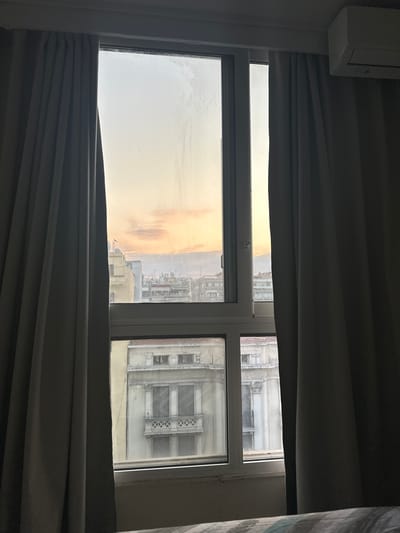 The image captures a view through a window, looking out at a cityscape. The window is framed by dark gray curtains, which are partially drawn to the sides, allowing a view of the exterior. The window itself is divided into sections, with the upper section revealing a sky at sunset or sunrise, displaying soft hues of orange, pink, and yellow. Below this, a lower section of the window offers a view of buildings, appearing to be in an urban setting.

The buildings visible are of varying architectural styles and ages. In the upper part of the window, there are several multi-story buildings with balconies and rooftops, some of which appear to be older structures. In the lower part of the window, a detailed view of an older, ornate building with classical architectural elements is visible, featuring columns, decorative facades, and balconies with intricate railings. The overall impression is of an established city with a mix of architectural heritage.

An air conditioning unit is mounted high on the wall to the right of the window. The scene appears to be from an indoor vantage point, likely a hotel room or apartment, as indicated by the presence of the air conditioner and the partially visible edge of a bed with patterned bedding in the foreground. The window panes have some smudges and streaks, suggesting they may not be perfectly clean.

While the image provides a detailed view of the urban environment and the architecture, there are no people, animals, or any discernible actions taking place in the scene. The focus is purely on the static view from the window.

Based on the architectural style of the buildings, particularly the ornate classical facade visible in the lower window section, and the general urban landscape, the location is consistent with areas in the Thessaloniki Municipal Unit, Greece, known for its historical architecture.