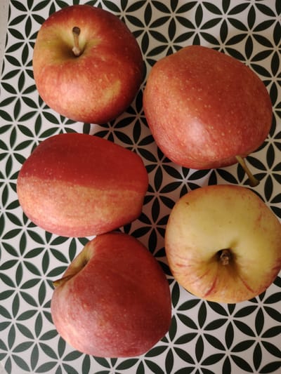Five red and yellow apples are arranged on a patterned surface. The surface has a repeating design of interlocking green leaf shapes on a white background. The apples vary in ripeness and color, with some exhibiting more red and others more yellow. One apple in the bottom right is cut in half, revealing its pale yellow flesh. There is no indication of a city or location within the image itself, and therefore no information can be provided about Кривец, Russia.