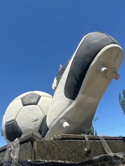 A monumental sculpture of a soccer ball and a cleat is displayed outdoors under a clear blue sky. The soccer ball, rendered in white with black pentagons, exhibits some visible cracks and wear. Adjacent to it, a large cleat sculpture, predominantly white with black accents on the upper portion and sole, is positioned with its studs visible. The sculptures rest on a granite base, which is partially covered with dark plastic sheeting and some transparent tape. In the background, trees and power lines are discernible against the sky. The location is identified as Lobacheve, Ukraine.