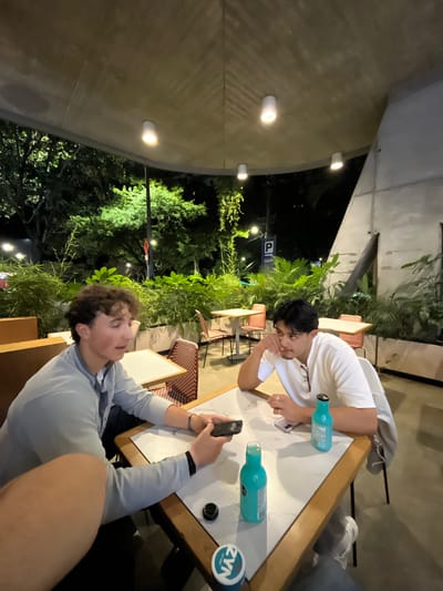 Two young men are sitting at a table outside, likely at a restaurant or cafe in Medellín, Colombia, based on the ambient lighting and vegetation. The man on the right is holding a smartphone, possibly taking a photo or using it. The other man in a white shirt is seated across from him, with a hand near his face in a pensive pose. Two blue bottles are on the table, along with a can with the word "ZYN" on it. Green foliage is visible in the background, suggesting an outdoor setting with plants. Additional tables and chairs of other diners are visible. The scene is illuminated by artificial lights affixed to a wall. The overall atmosphere suggests a casual, relaxed gathering in an urban environment.