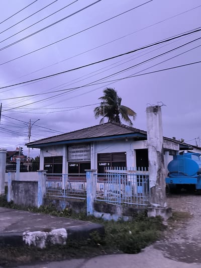 An eye-level shot captures a weathered public utility building under an overcast, purplish-grey sky, likely in Jurong Peukan, Indonesia. Numerous power lines crisscross the sky above. The single-story building features a dark, corrugated roof and light-colored walls, appearing faded and stained. A prominent sign on the front of the building, written in Indonesian, reads "PEMERINTAH KABUPATEN PIDIE" (Government of Pidie Regency), "PERUSAHAAN UMUM DAERAH AIR MINUM" (Regional Drinking Water Company), and "TIRTA MON KRUENG BARO." Below the main text, signs for "LOKET 1," "LOKET 2," and "LOKET 3" indicate service windows.

A blue metal fence with concrete posts, showing signs of wear and peeling paint, runs along the front of the building. Overgrown grass and weeds are visible along the base of the fence and in the foreground, where a concrete curb or path is present. Behind the building, a tall palm tree with coconuts is visible, its fronds appearing wind-swept. To the right of the building, a large, blue water tank truck is partially visible, parked behind a tall, weathered concrete pillar. The overall scene suggests a late afternoon or early evening ambiance, with subdued lighting.