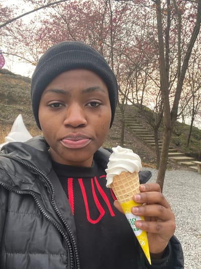An individual with dark skin, wearing a black beanie, a black puffer jacket, and a black sweater with red lettering, is centered in the frame. The person looks directly forward, with their tongue slightly extended between their lips. In their right hand, they hold a single-scoop vanilla soft-serve ice cream cone, encased in a yellow paper sleeve featuring Chinese characters. To the left, a large white sculpture resembling a swirl of soft-serve ice cream is partially visible on an earthen slope. The background reveals a sloping terrain covered in light-colored gravel or small stones, with a set of concrete or stone steps ascending the hillside to the right. Numerous deciduous trees with sparse branches and clusters of pink blossoms or leaves are scattered across the background against a light sky. The scene is located outdoors in Caijia, China.