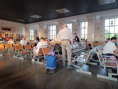 This is an eye-level, wide shot taken inside the Orsha, Belarus train station waiting area. The room is large with a dark, tiled ceiling and several large, classical-style columns supporting it. Natural light streams in from large windows lining the far wall. The floor is a speckled dark gray tile. Rows of orange, wooden bench seats with metal frames are arranged throughout the space. Many people are seated, waiting, with various bags and luggage near them. In the foreground, a man in a plaid shirt and tan pants is walking, and a blue rolling shopping bag with a floral pattern is parked next to a seat. A backpack is on the seat behind him. To the left, another man is standing and talking to a seated woman. Further into the waiting area, more people are seated or walking. The atmosphere appears calm and functional, typical of a transportation hub.