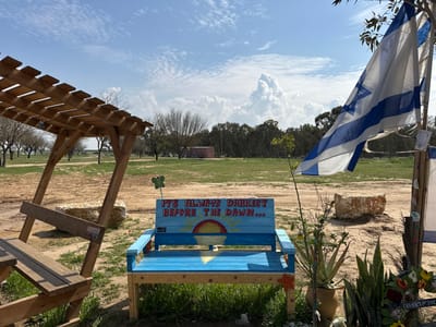 The image shows a wooden bench painted blue with the inscription "IT'S ALWAYS DARKEST BEFORE THE DAWN..." and a large, white rock on the ground nearby. The bench is located in an open, grassy field with a wooden pergola in the foreground and an Israeli flag waving in the wind in the background. The sky is blue with white fluffy clouds.  The environment is dry and dusty with some trees in the distance. The location, Re'im, Israel, is a small, rural community in the Negev desert. The coordinates, 31.398 latitude and 34.471 longitude, pinpoint the location within Re'im.
