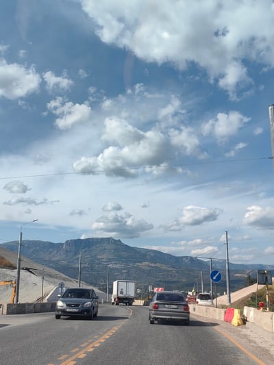 The image shows a view from a vehicle on a highway, looking forward towards a mountainous landscape under a partly cloudy sky. Several vehicles are visible on the road, including a dark gray Kia Rio in the foreground with the license plate B053KA 92. Behind it, a white truck and another car are traveling in the same direction. To the right of the road, there are construction barriers, traffic cones, and signs, including a blue circular sign with a white arrow indicating a left turn. The setting appears to be a sunny day, possibly late morning or early afternoon, given the bright sunlight and distinct shadows. The mountains in the background are rugged and appear to be covered in vegetation and some scattered buildings. The road itself has double yellow lines and lane markers. The overall scene depicts a common highway journey with a scenic backdrop.