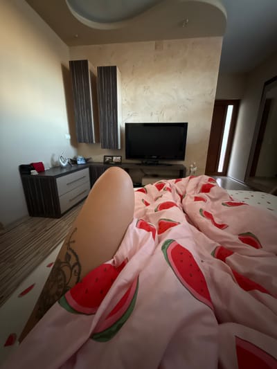 The image depicts an interior scene, likely a bedroom, within a residence in Rakovski, Bulgaria. In the foreground, a person's leg, adorned with a tattoo, is visible, partially covered by a pink duvet featuring a watermelon pattern. The duvet is spread across a bed. Adjacent to the bed, to the left, is a dark wooden dresser with two drawers and a smaller, darker cabinet on top, which holds a red item, possibly a bag, and a silver decorative object. Further to the left, a wall with a textured finish is visible. In the background, a black television set is mounted on a stand. Above the television, two tall, dark wooden cabinets with vertical striped detailing are mounted on the wall. The overall environment suggests a residential setting.