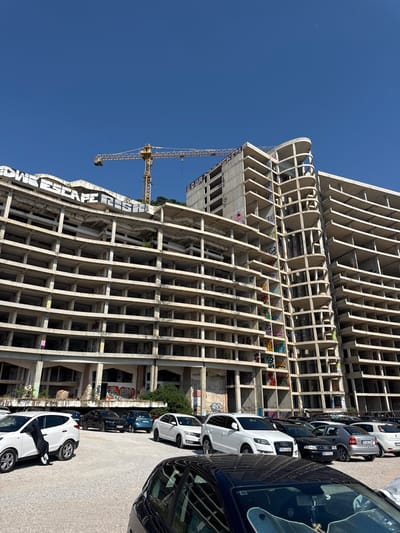 The image shows an unfinished concrete building with a crane in the background. The building has a skeletal structure with exposed floors and support beams. Graffiti covers some of the concrete surfaces. In front of the building, a row of cars is parked. The scene is set on a bright, sunny day under a clear blue sky. The time of day appears to be daytime. The location is Perazića Do, Montenegro, as indicated by the text "Escape" and "Perazića Do" visible on the building. The overall atmosphere is one of urban decay and abandonment, with the unfinished construction juxtaposed against the parked vehicles.