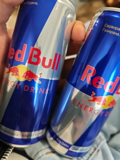The image shows two cans of Red Bull energy drink. The cans are blue and silver with the Red Bull logo prominently displayed. The text on the cans is in Bulgarian. The cans are likely located in Grohotno, Bulgaria based on the Bulgarian text. A hand is holding one of the cans. The cans appear to be resting on a pair of jeans.